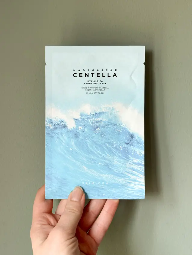 Hand holding a SKIN1004 Madagascar Centella Hyalu-Cica Hydrating Mask packet against a sage green wall.