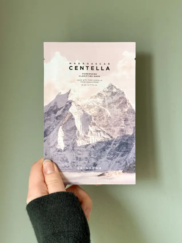 Hand holding a SKIN1004 Madagascar Centella Poremizing Clarifying Mask packet against a sage green wall.
