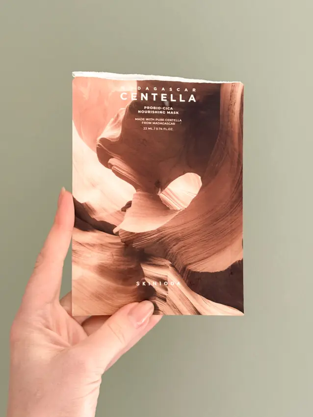 Hand holding an open SKIN1004 Madagascar Centella Probio-Cica Nourishing Mask packet against a sage green wall.