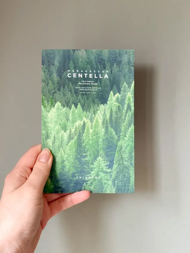 Hand holding a SKIN1004 Madagascar Centella Tea-Trica Relaxing Mask packet against a sage green wall.