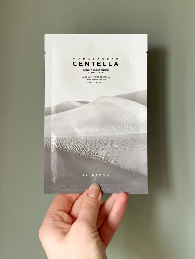 Hand holding a SKIN1004 Madagascar Centella Tone Brightening Glow Mask packet against a sage green wall.