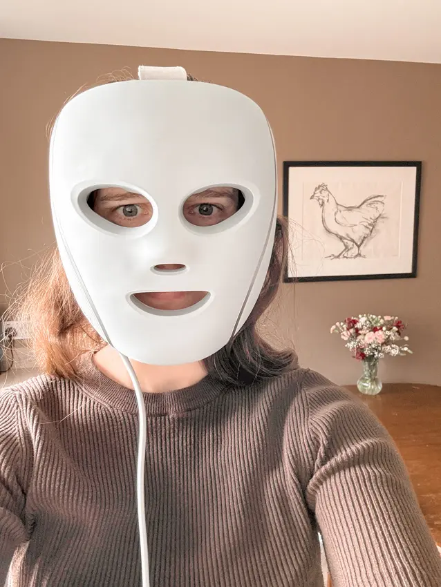 Wearing the grey Shark CryoGlow LED face mask with its top strap fitted, standing against a brown wall.