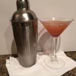 A pink blackberry cocktail in a martini glass next to a stainless steel cocktail shaker on a white cloth