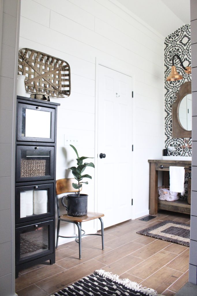 Modern farmhouse bathroom with white shiplap walls, industrial glass front storage cabinet, vintage school chair, and wood look tile flooring