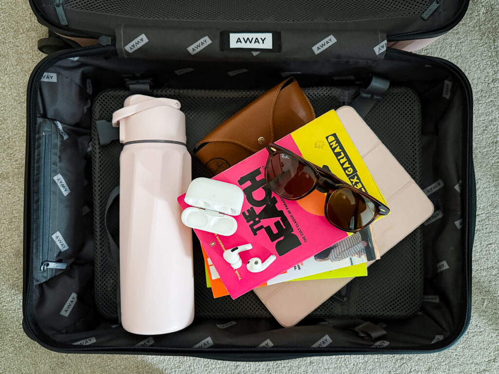 carry-on away case packed with books, ray ban sunglasses, a water bottle, airpods and an ipad pro