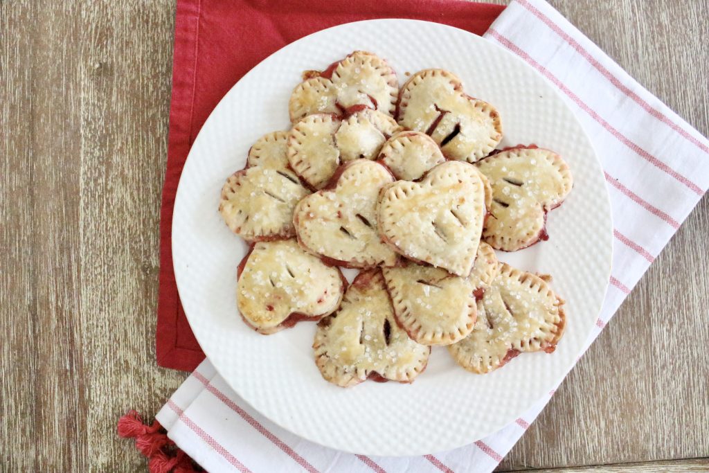 A plate of baked heart shaped hand pies with fork crimped edges and sparkling sugar seen from above on a red napkin