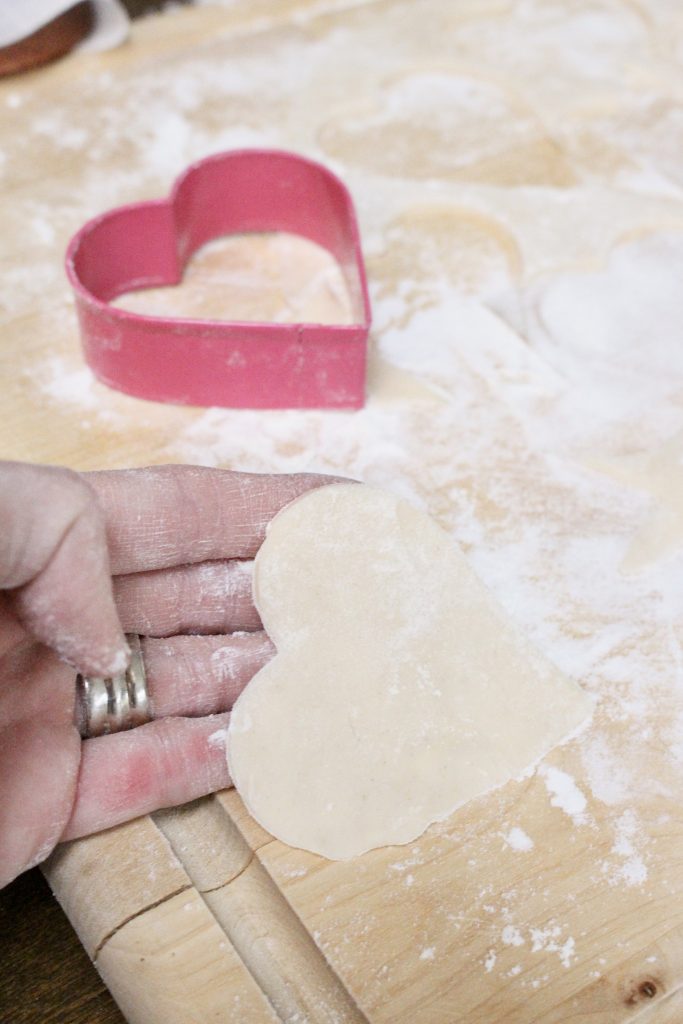 A hand holding a small heart shaped piece of pastry cut from rolled pie crust on a floured wooden board