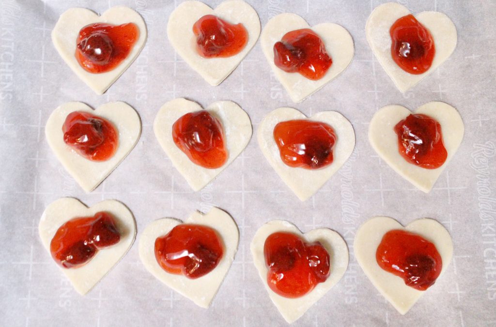 Heart shaped pastry bases filled with strawberry jam on parchment paper before adding the pastry lids