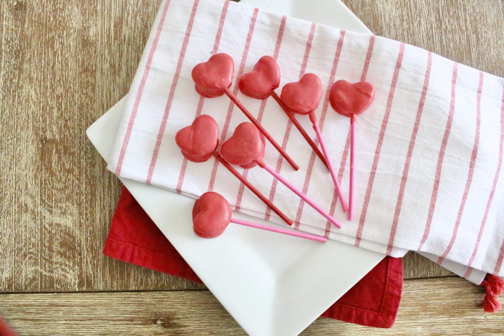 Pink chocolate coated heart marshmallow pops arranged on a white plate with a striped tea towel and red napkin