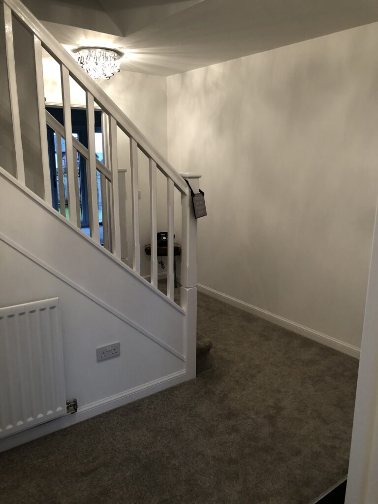 Hallway before the makeover showing plain white walls, grey carpet, crystal ceiling light and radiator under the stairs