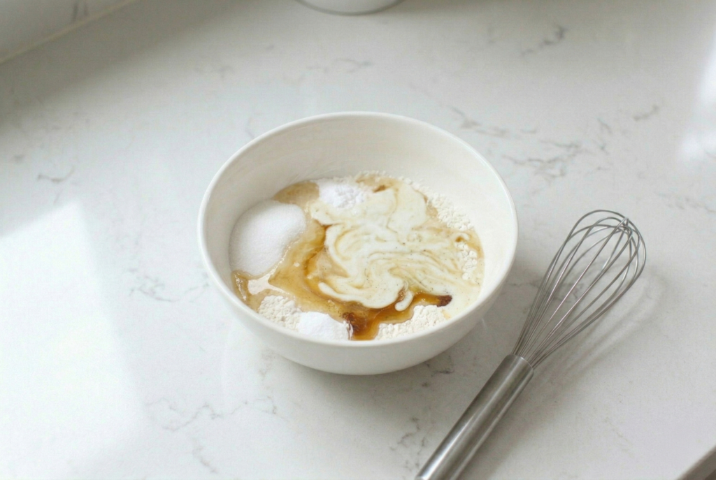 Dry pancake batter ingredients in a white ceramic bowl with vanilla extract and oil added, next to a silver whisk on a marble worktop