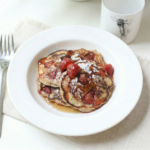 Fluffy vegan strawberry pancakes stacked on a white plate, topped with fresh strawberries, maple syrup and a dusting of icing sugar