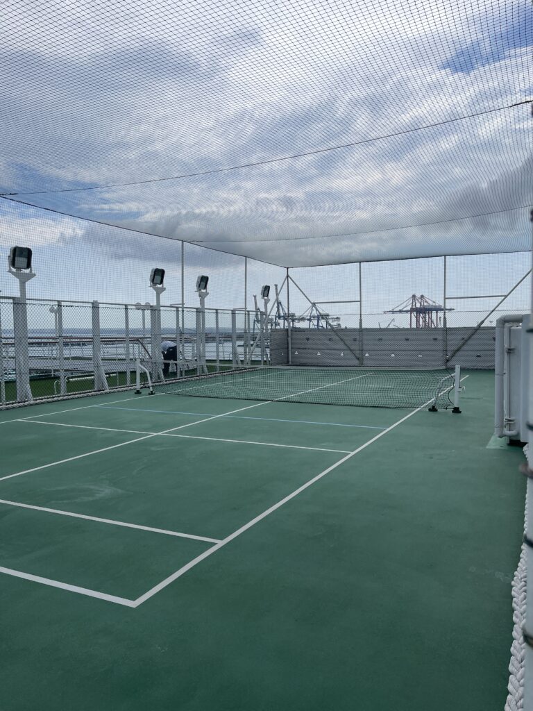 The outdoor tennis court on Crystal Symphony cruise ship, surrounded by protective netting and overlooking the ocean and dockyard cranes under a partly cloudy sky.