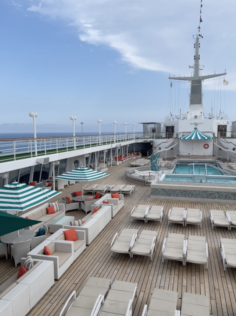 The pool deck of Crystal Symphony cruise ship, featuring striped green-and-white umbrellas, cozy seating with orange cushions, sun loungers, and a serene swimming pool under a clear blue sky.