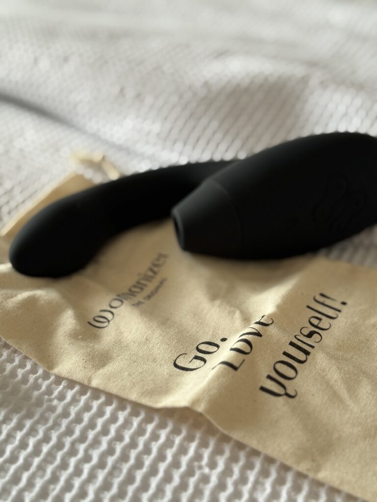 Womanizer Duo 2 laying on a cream cotton pouch with the text "Go. Love yourself!" on a soft white bedspread.