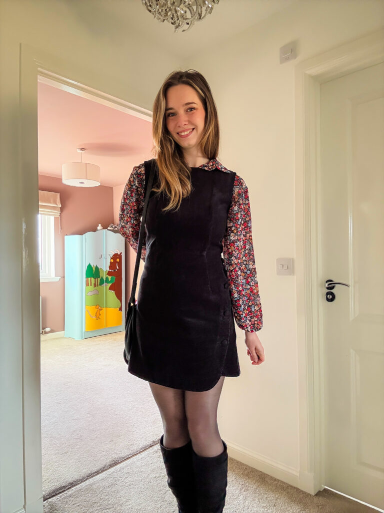 Layered autumn outfit with a floral blouse under a black pinafore dress, styled with black tights and ankle boots.