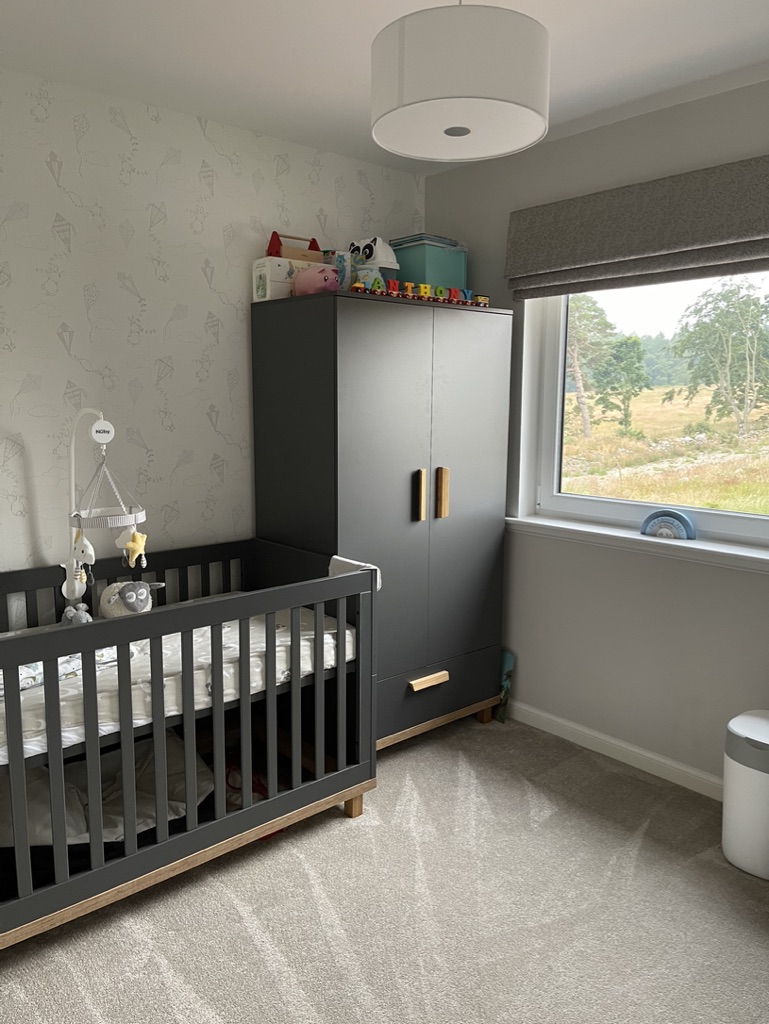 Minimalist nursery with grey cot, black wardrobe with wooden handles, and soft kite-print wallpaper beside a large window.