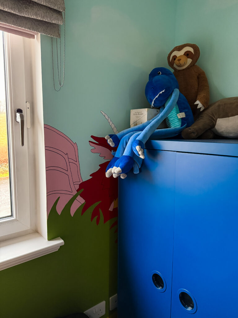 Painted dragon mural partially hidden behind a blue wardrobe in a child’s room with cuddly toys on top.