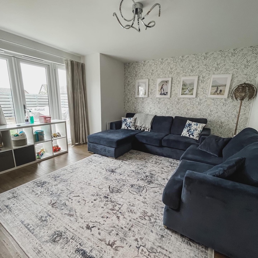Before image of living room featuring navy sectional sofa, blue-patterned rug, floral wallpaper, and framed family photos above the sofa.