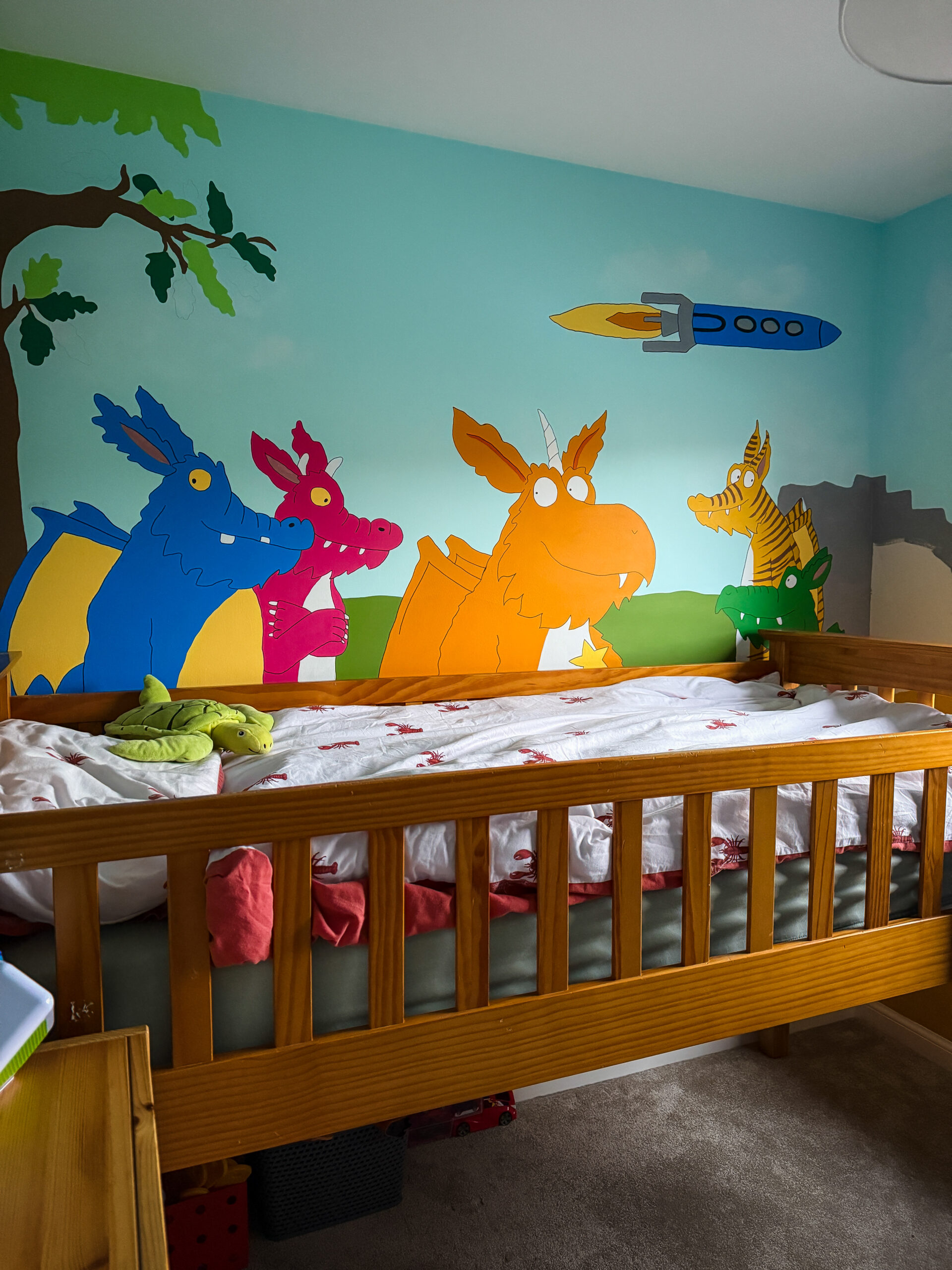 Colourful mural of Zog and dragon friends painted on a bedroom wall, mostly hidden behind a mid-height wooden bed.