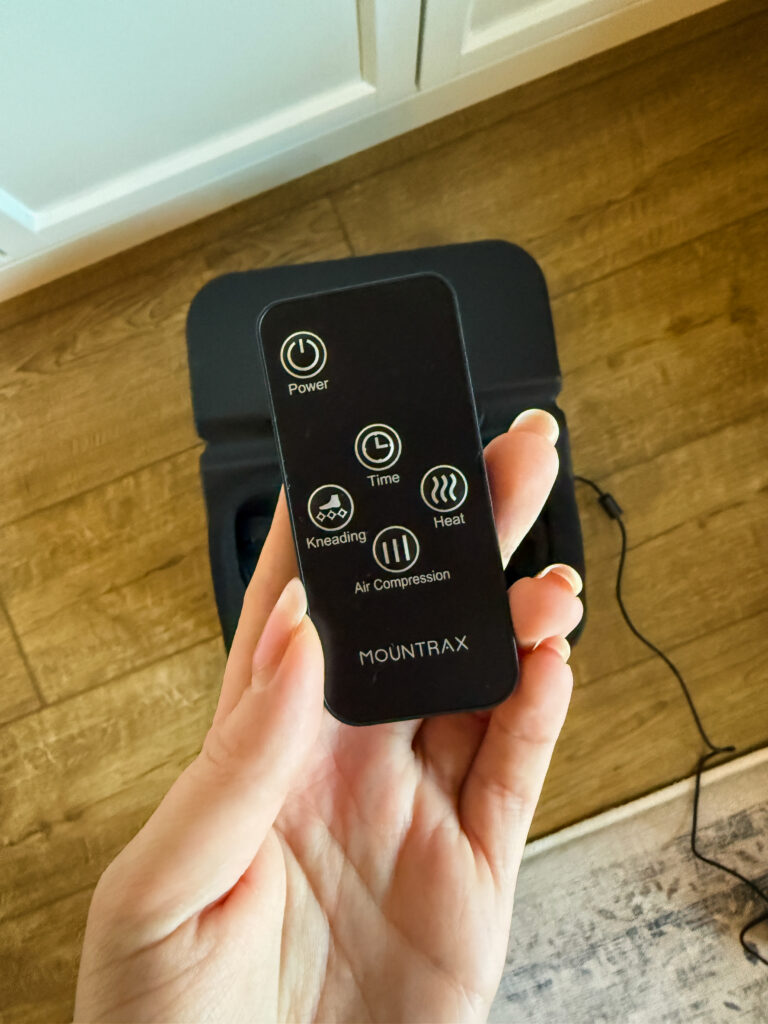 Hand holding MOUNTRAX foot massager remote control showing power, time, heat, kneading and air compression buttons