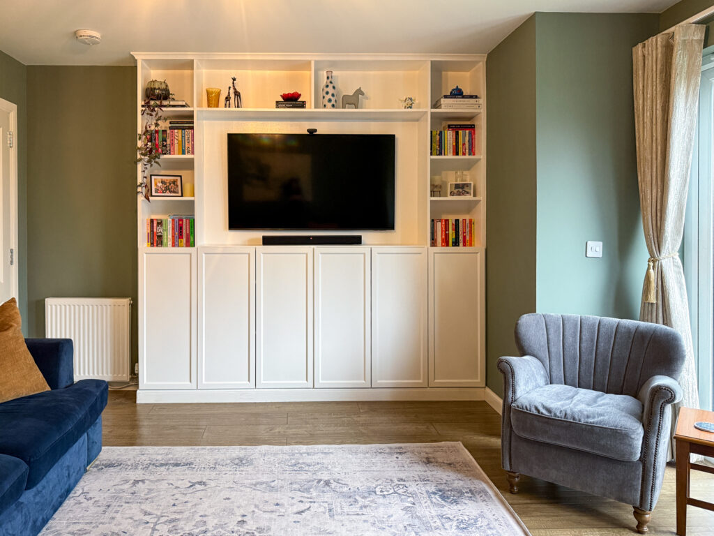 Full view of white Billy bookcase built-ins with wall-mounted TV and grey velvet armchair against Farrow and Ball Card Room Green painted walls