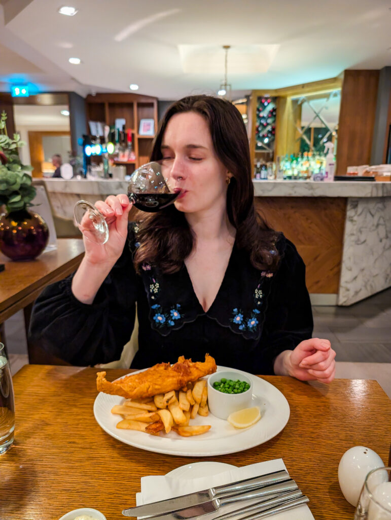 A woman holding a glass of red wine at dinner in the Craigendarroch hotel restaurant, with a plate of fish and chips in front of her