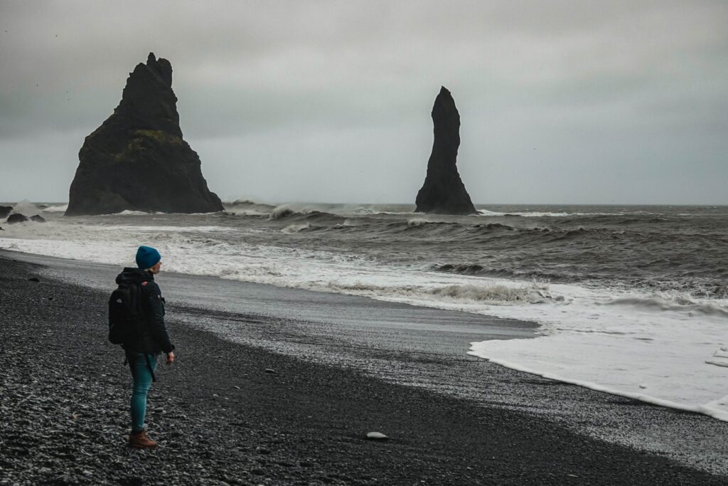 Traveller wearing winter gear and blue beanie standing on the black-sand beach at Vík in Iceland facing dramatic sea stacks in misty weather.