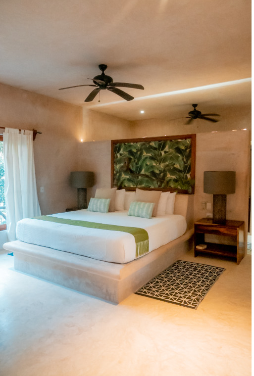 Spacious villa bedroom at Cachito de Cielo Tulum, with a large white bed, tropical leaf artwork as a headboard, ceiling fans, and soft neutral plaster walls