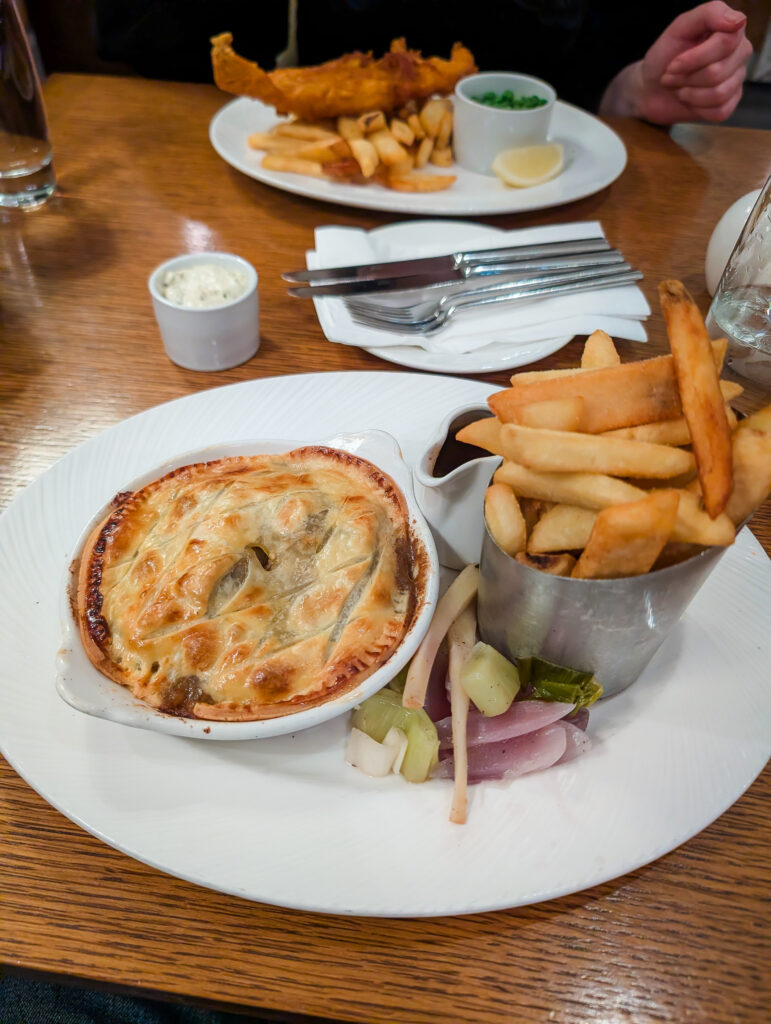 A steak pie in a ceramic dish served with chips and vegetables at the Craigendarroch hotel restaurant, with a plate of fish and chips visible in the background