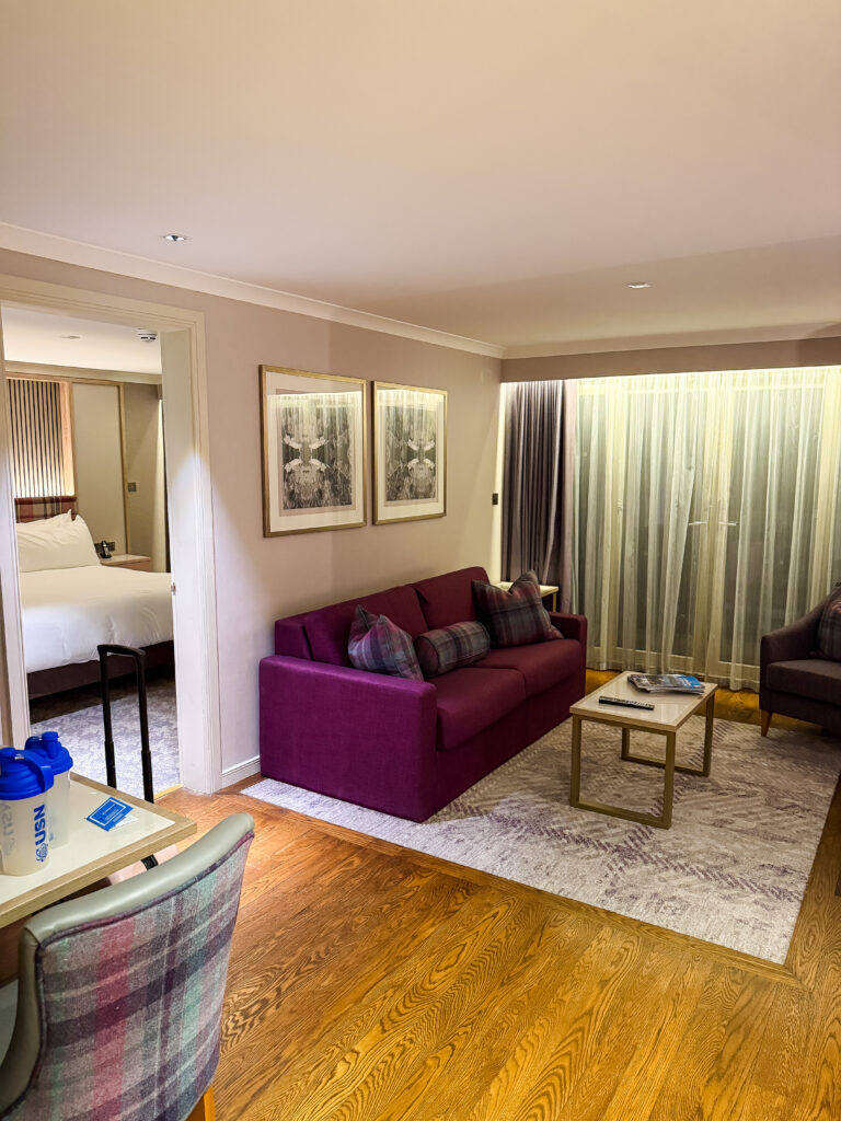 The spacious lounge area of a Craigendarroch Hilton Suite, with a deep burgundy sofa, framed artwork on the walls, and warm parquet flooring