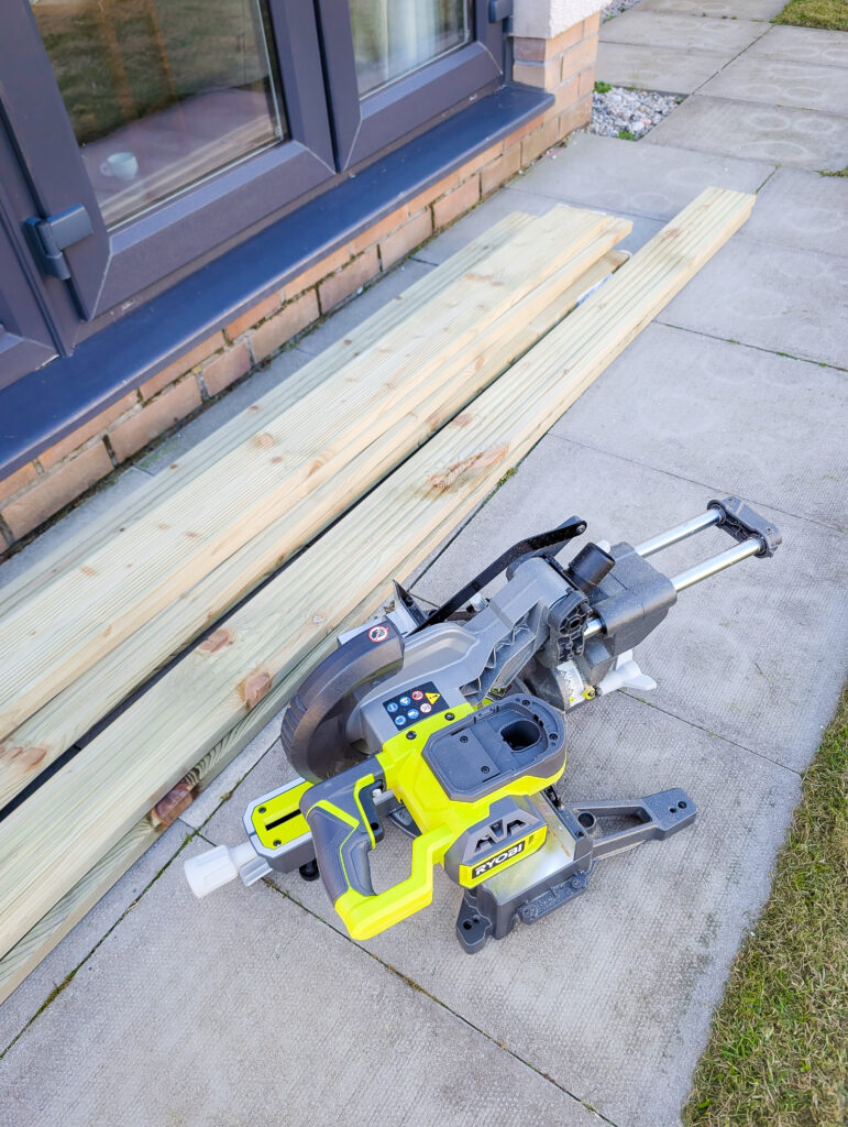 Treated decking boards laid out alongside a Ryobi mitre saw ready to be cut to size for the raised vegetable planter