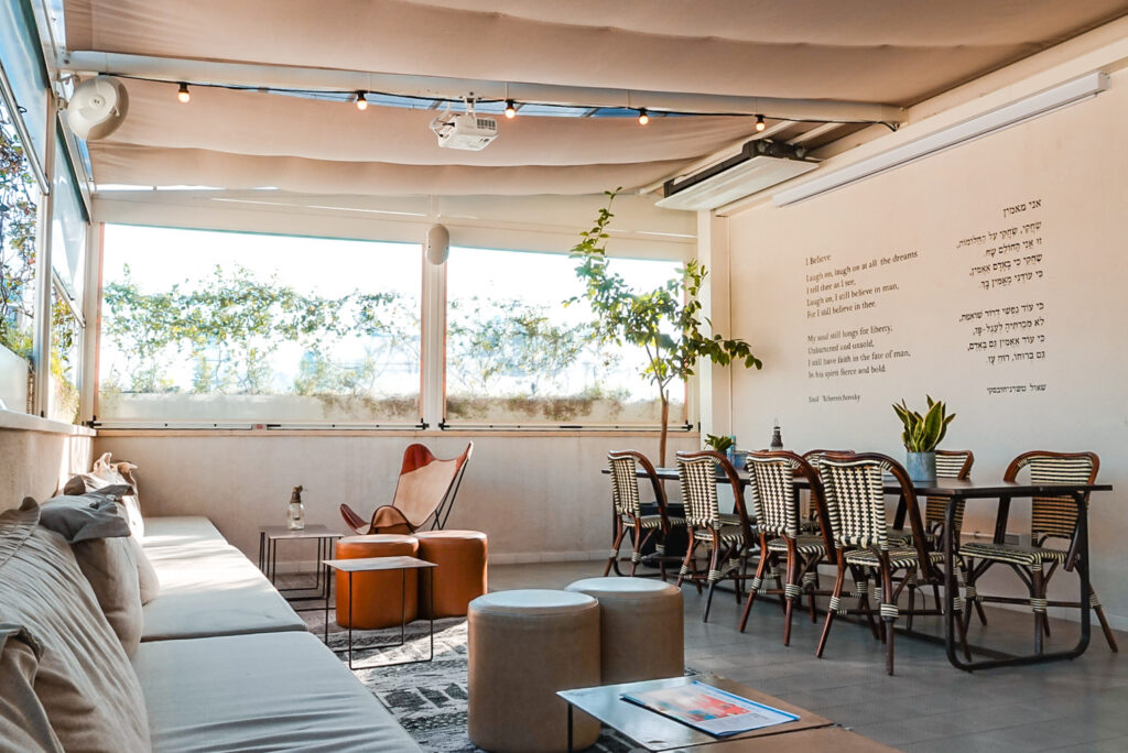 The bright covered terrace lounge at Hotel Saul Tel Aviv, with sofas, an armchair, dining table, and string lights overhead