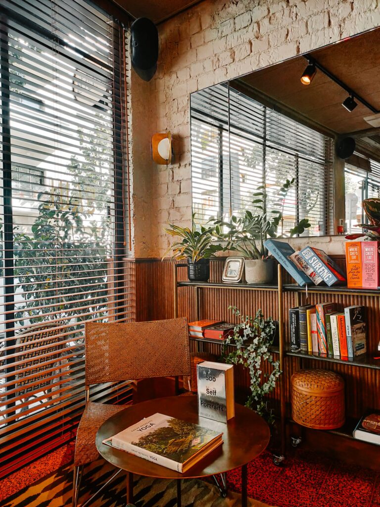 A cosy reading corner in the Hotel Saul Tel Aviv lobby, with a rattan chair, a small coffee table with books, and a bookshelf beside a window with wooden blinds