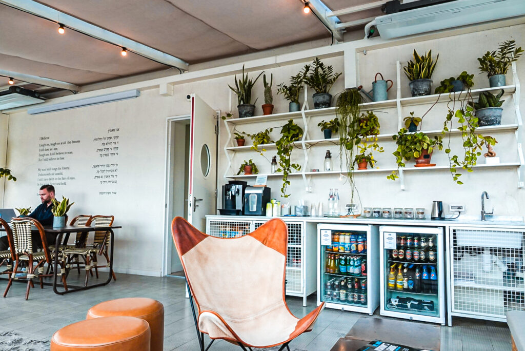 The self-serve drinks station on the Hotel Saul Tel Aviv terrace, with a bar fridge stocked with cold drinks, coffee machine, and plant-lined shelves above