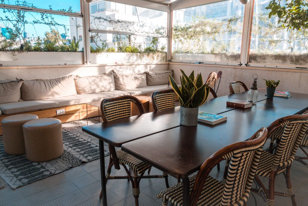 Relaxed seating area on the covered terrace at Hotel Saul Tel Aviv, with banquette cushions, wicker chairs, and city views through the windows