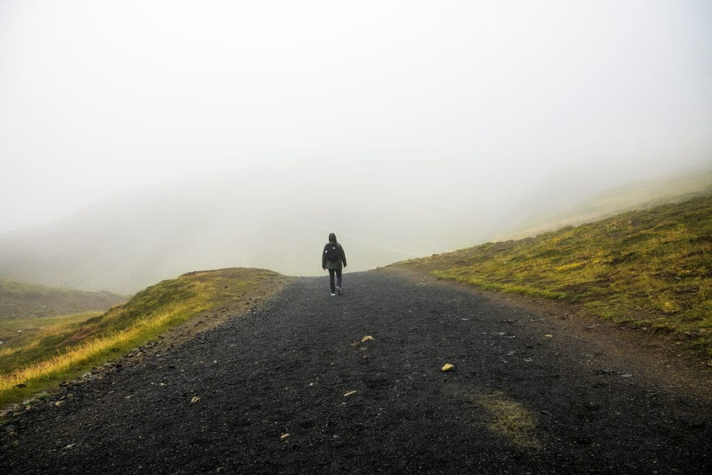 Lone hiker walking into thick mist along a gravel path through green Icelandic hills
