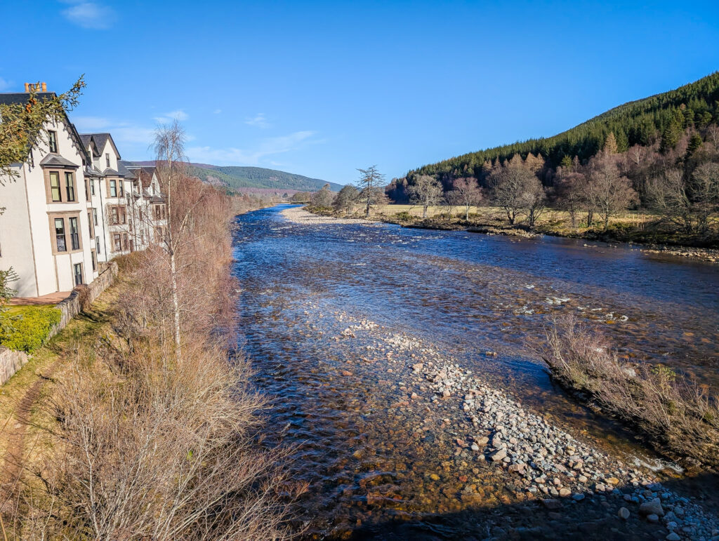The River Dee flowing through Ballater in Royal Deeside on a clear sunny day, with white-painted buildings on the bank and wooded hills beyond
