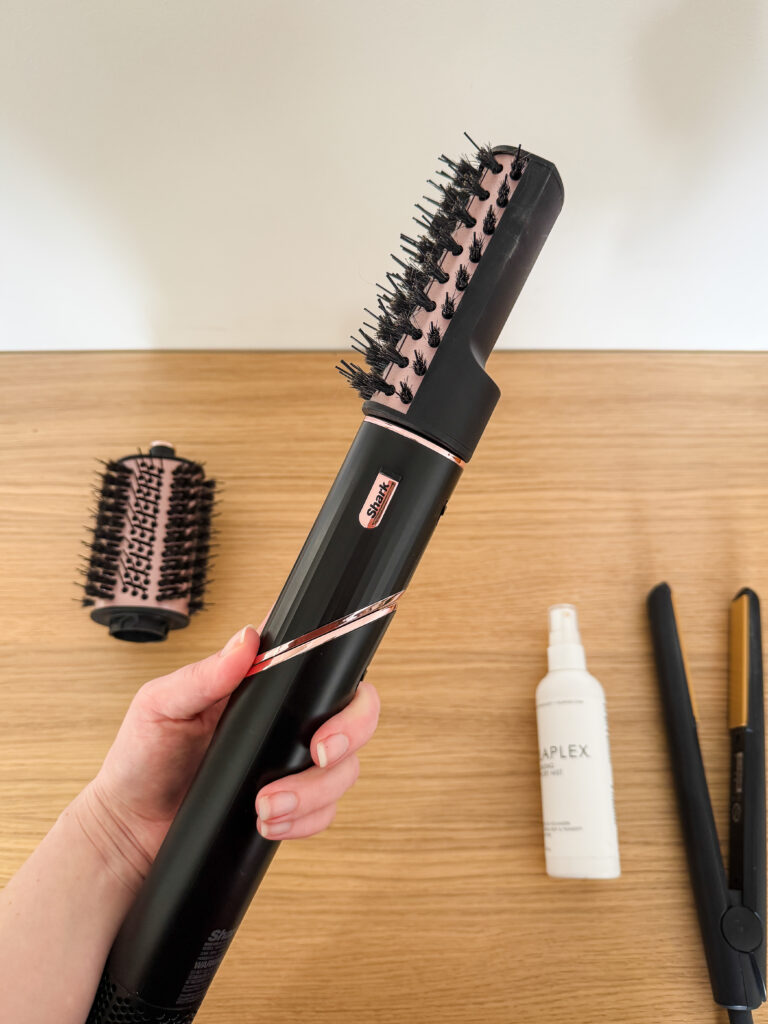 A hand holding the black and rose gold Shark FlexStyle multi-styler with the smoothing paddle brush attachment connected.