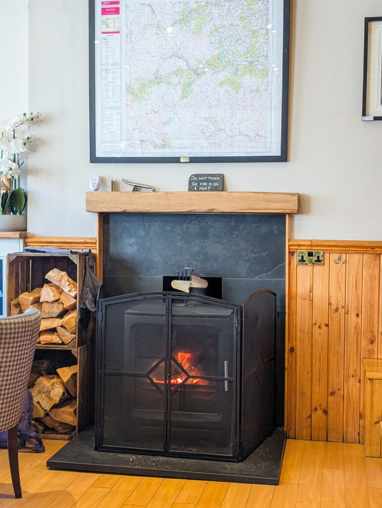 A lit log-burning stove inside The Bothy café in Ballater, with stacked firewood to the side and a framed Ordnance Survey map of the area on the wall above