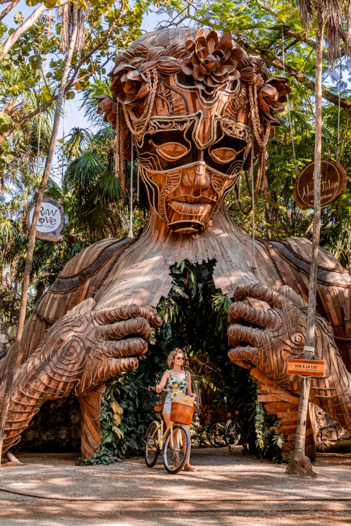 Woman with bicycle standing beneath a giant wooden Aztec-style statue on the Tulum hotel road