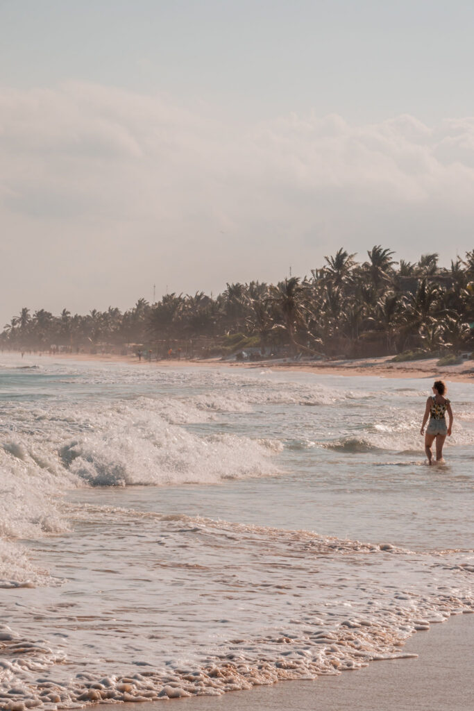 Woman walking along the shoreline at Tulum beach at sunset, palm trees lining the coast