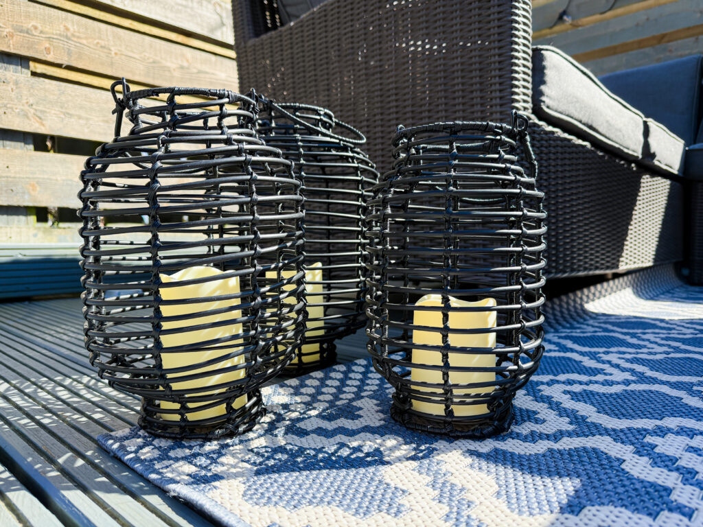 Three black woven rattan lanterns with ivory LED pillar candles sitting on a blue outdoor rug.