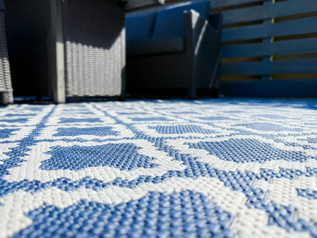 A close-up, low-angle shot showing the texture of a blue and white geometric patterned outdoor rug on grey decking.