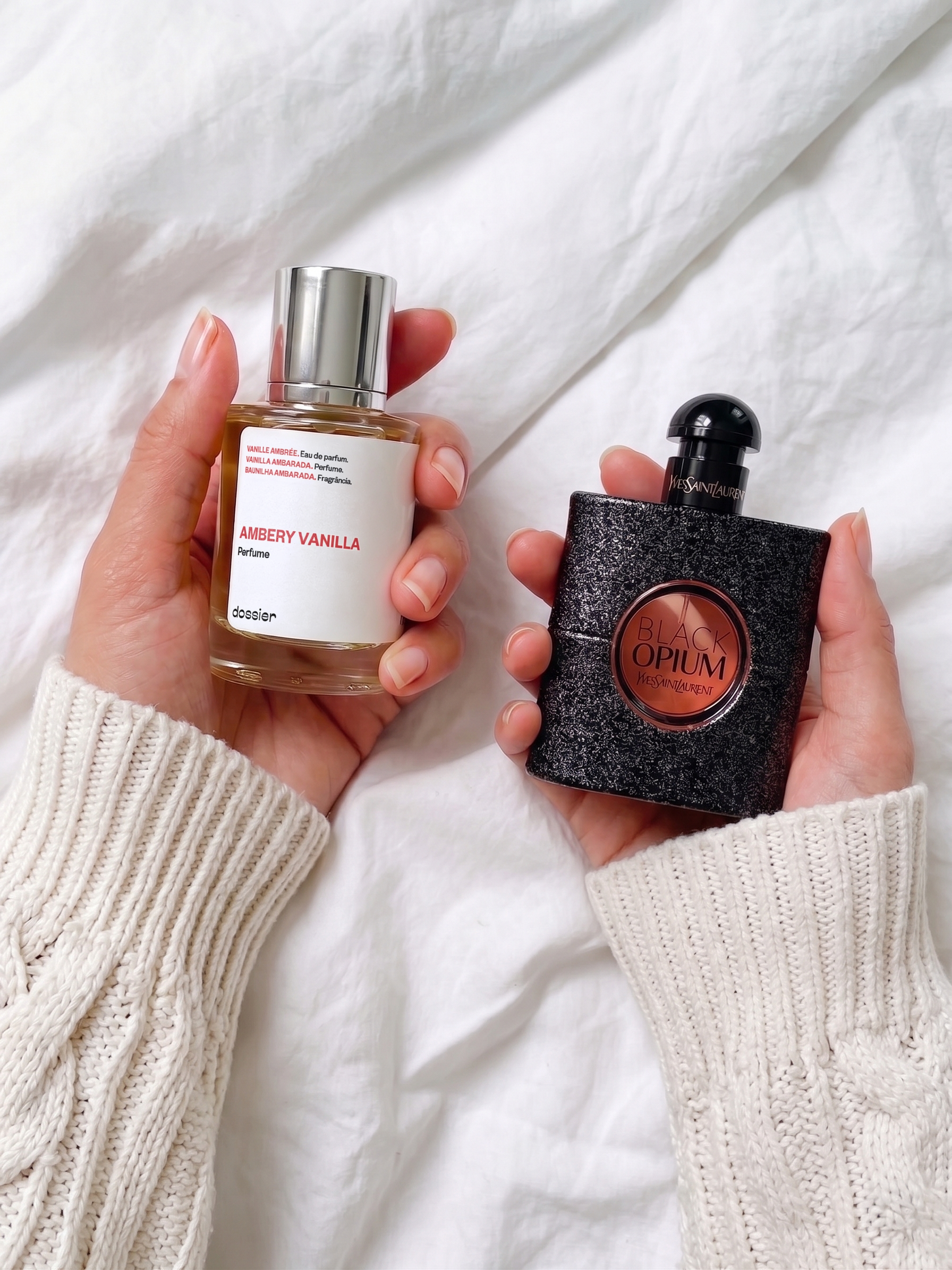 A person wearing a white cable-knit sweater holding a bottle of Dossier Ambery Vanilla in one hand and YSL Black Opium in the other against a white linen background.