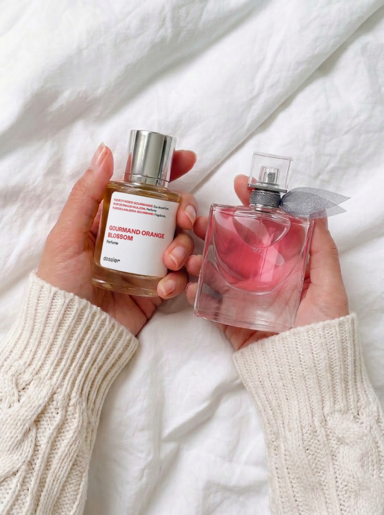 A person wearing a white cable-knit sweater holding a bottle of Dossier Gourmand Orange Blossom in one hand and Lancome La Vie Est Belle in the other against a white linen background.