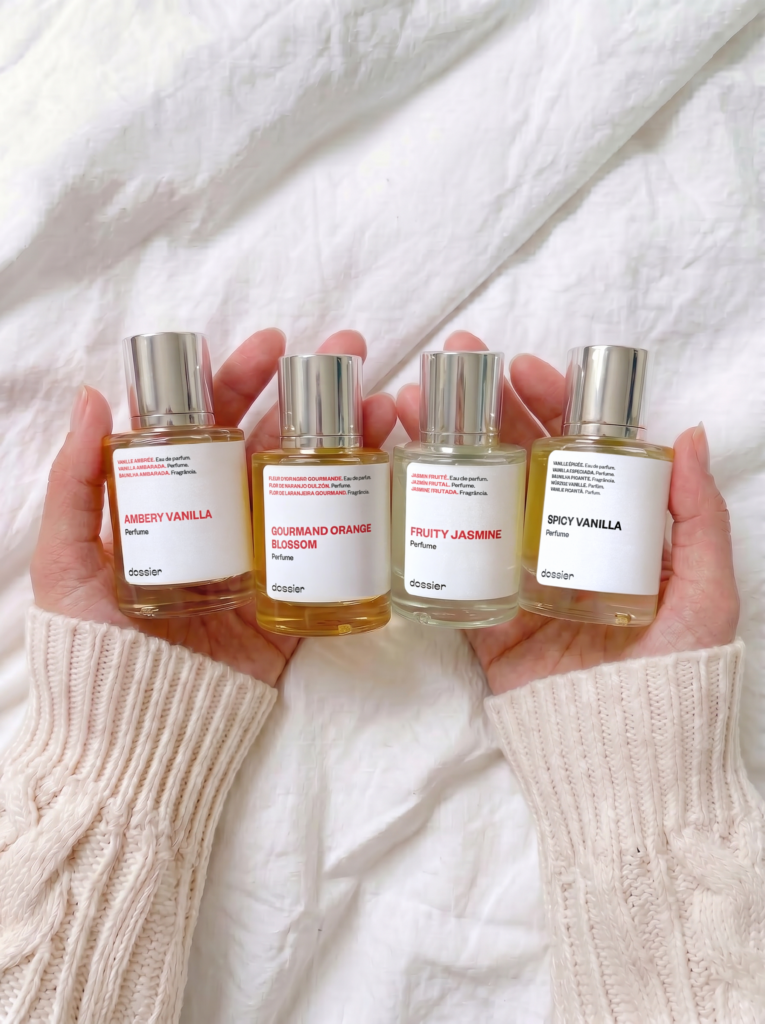 A person wearing a cream cable-knit sweater holding four Dossier perfume bottles: Ambery Vanilla, Gourmand Orange Blossom, Fruity Jasmine, and Spicy Vanilla. The bottles are lined up in their hands against a soft white linen background.