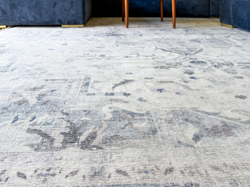 A large washable Ruggable rug tucked under a navy blue velvet sofa, demonstrating how a 275 x 365cm rug fits in a standard living space.