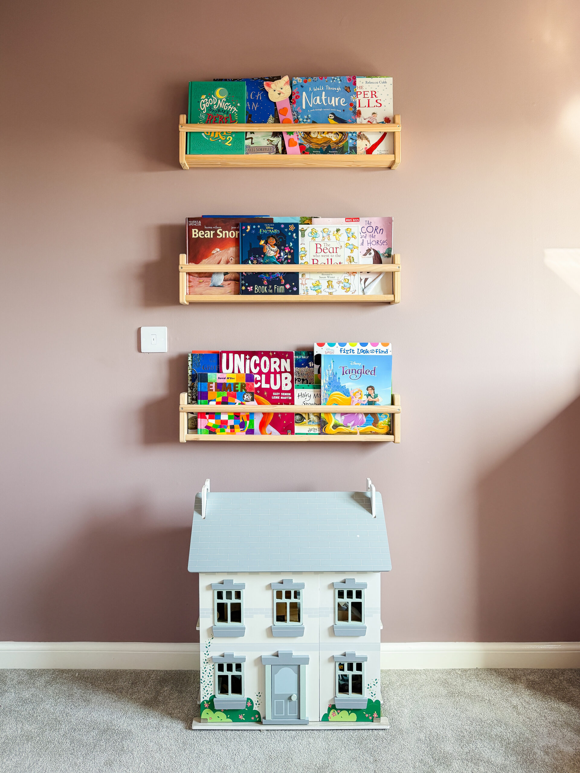 wooden book display shelves filled with children's books above a white dollhouse against farrow and ball sulking room pink walls