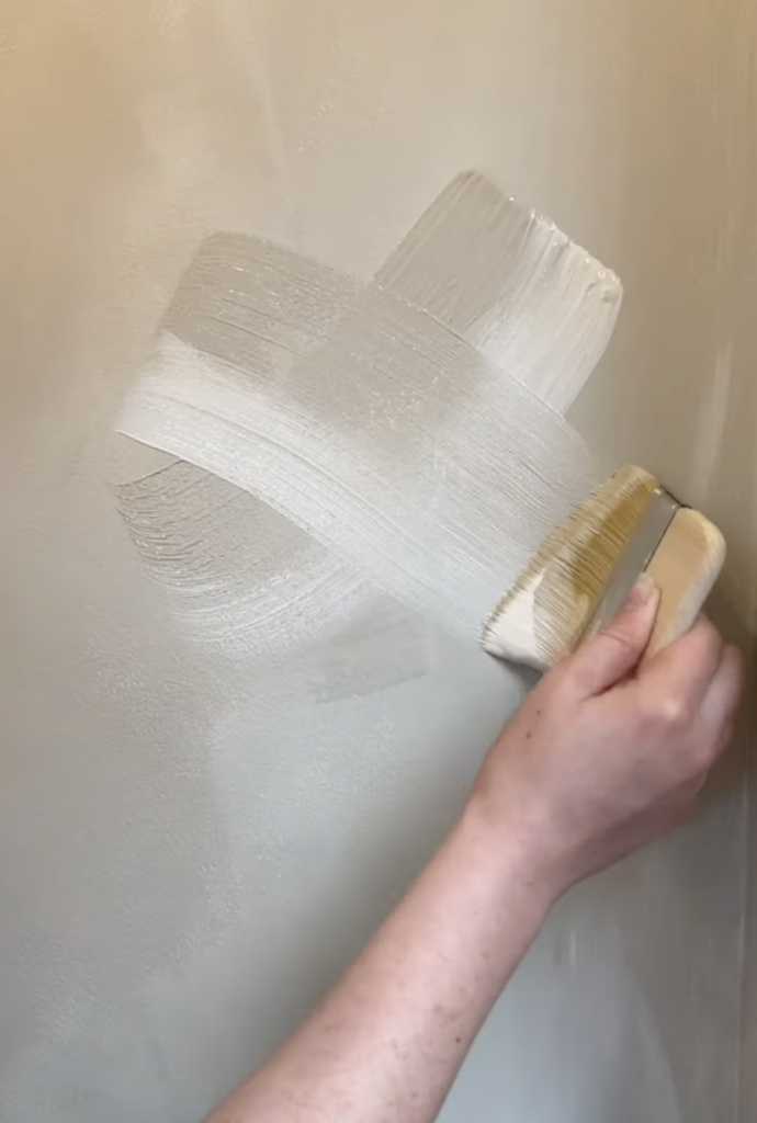 hand applying faux limewash paint to a wall using a wide brush in cross strokes